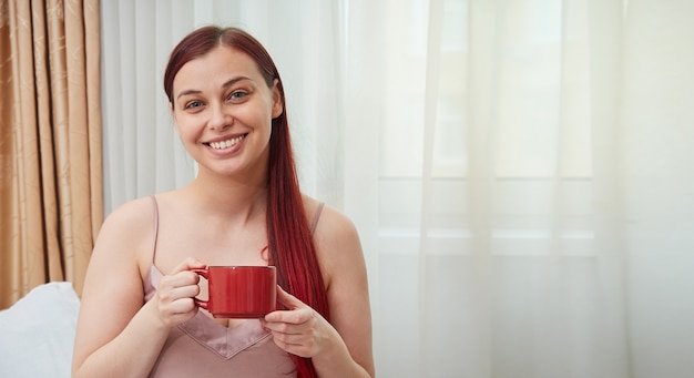 Vrouw met rood haar zit op de achtergrond van het raam met een kopje koffie in de ochtend met kopieerruimte