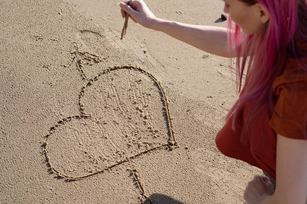 vrouw met roze haar tekent een hart met een stok op het zand