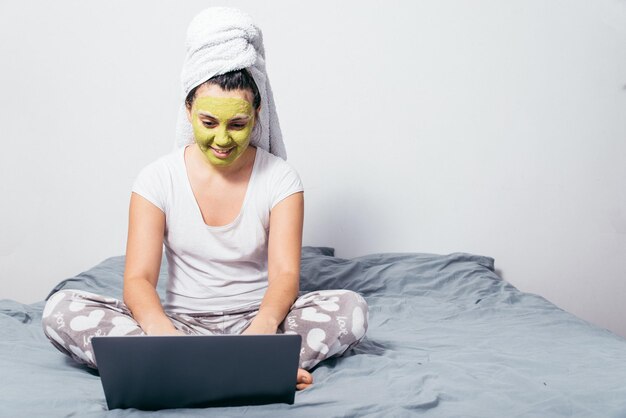 Vrouw met schoonheidsmasker op gezicht zit op bed met laptop