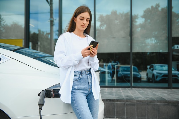 Vrouw met telefoon in de buurt van een elektrische huurauto Voertuig opgeladen bij het laadstation