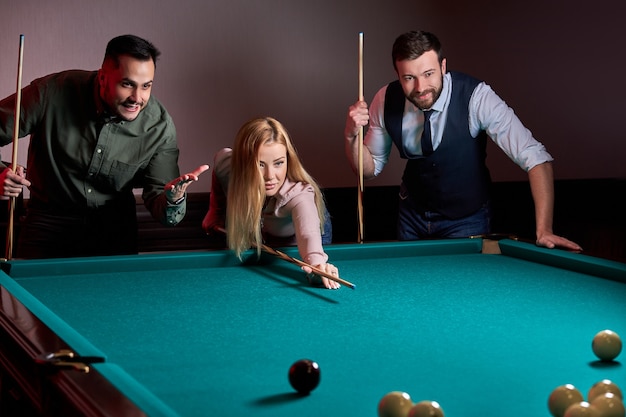 Vrouw met twee mannelijke vrienden spelen biljart in de bar na het werk, hebben rust en vrije tijd en bereiden zich voor om poolballen te schieten