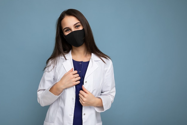 Vrouw met virusbeschermend masker op gezicht tegen coronavirus en witte medische jas geïsoleerd op blauwe achtergrond. Ruimte kopiëren