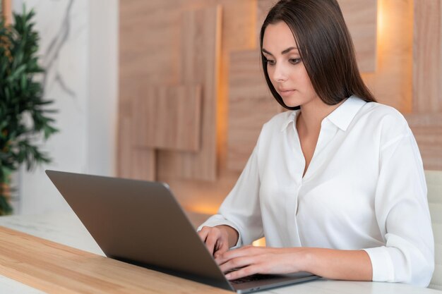 Vrouw met wit overhemd zittend aan tafel met laptop en notebook die aantekeningen maakt Concept van werk