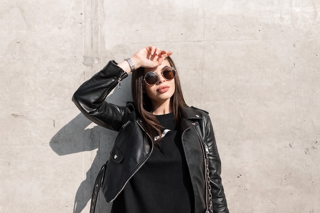 Vrouw model met natuurlijke schoonheid in vintage ronde zonnebril en modieuze zwarte kleding met leren jas staat in de buurt van een betonnen muur in de stad in zonlicht