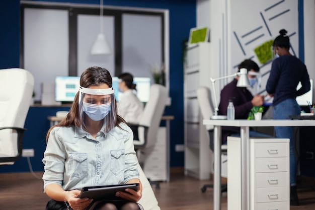 Vrouw ondernemer met beschermingsmasker tegen covid-19 pandemie zittend in het midden van de kantoorruimte met een nieuwe normale digitale tablet. Multi-etnisch zakelijk team dat werkt met respect voor sociale afstand