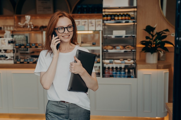 Vrouw ondernemer met rood haar heeft telefoongesprek houdt laptop vast