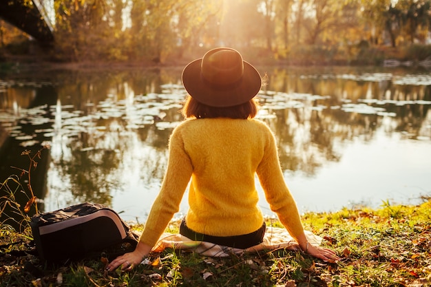 Vrouw ontspannen door herfst rivier bij zonsondergang. Meisjeszitting op bank door brug die van landschap geniet. herfstwandeling