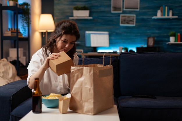 Vrouw op de bank die hamburgerdoos uitpakt uit een papieren afhaalzak zittend op de bank voor tafel met bierfles, friet en kom chips. Persoon die in de woonkamer fastfood thuis uitpakt.