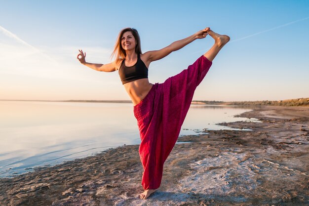 Vrouw op het strand bij zonsondergang begint yoga asana-training te doen. Ochtend natuurlijke stretch warming-up training