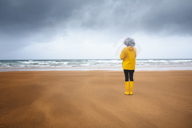 Vrouw op het strand van achteren gezien, kijkend naar de zee in de regen, met een transparante paraplu, gekleed in een regenjas en gele laarzen, op een bewolkte dag met stormen. kopieer ruimte