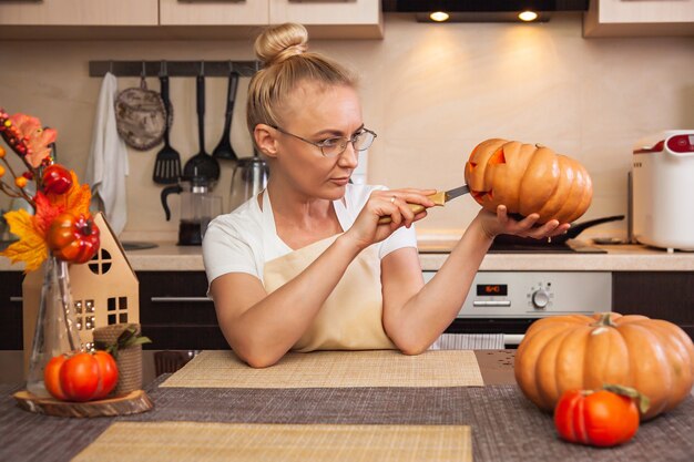 Vrouw op keuken snijdt een pompoen voor Halloween in een kamer met herfstdecor en een lamphuis. Gezellig huis en voorbereiding op Halloween.