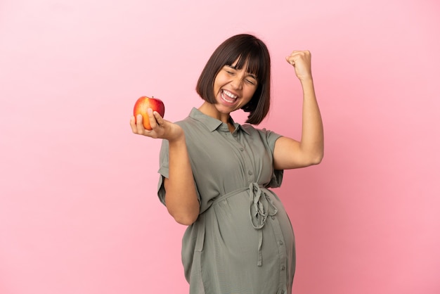 Vrouw over geïsoleerde achtergrond zwanger en houdt een appel vast terwijl ze een overwinning viert
