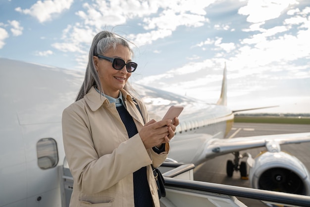 Vrouw passagier in zonnebril met een telefoon staat op de trappen van het vliegtuig voor het instappen