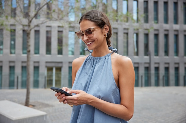 vrouw poseert in stedelijke omgeving tijdens zonnige dag draagt zonnebril blauwe jurk gebruikt smartphonegadget voor communicatiechats online gebruikt navigator om route te vinden