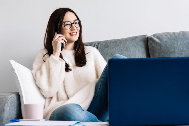 Vrouw praat met mobiele telefoon terwijl ze werkt met een laptop die thuis op een bank zit