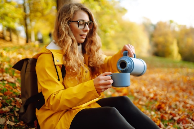 Vrouw reiziger in gele jas zit op een open plek tussen gele bladeren en drinkt warme drank uit thermoskan