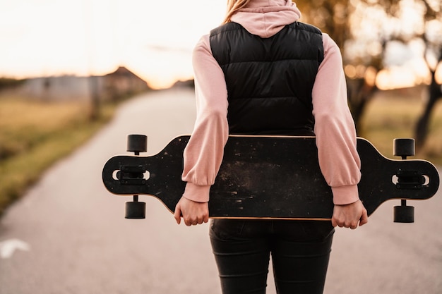 Vrouw rijdt op rechte weg op longboard bij zonsondergang Schaatser in vrijetijdskleding training aan boord tijdens avondzonsondergang met oranje licht Meisje houdt longboard in handen