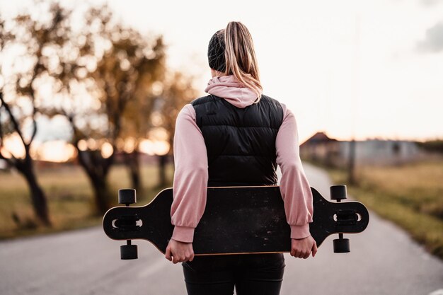 Vrouw rijdt op rechte weg op longboard bij zonsondergang Schaatser in vrijetijdskleding training aan boord tijdens avondzonsondergang met oranje licht Meisje houdt longboard in handen