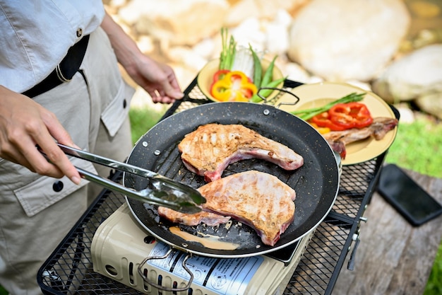 Vrouw roostert een varkenskotelet biefstuk op een vliegende pan koken met een draagbare picknick kachel