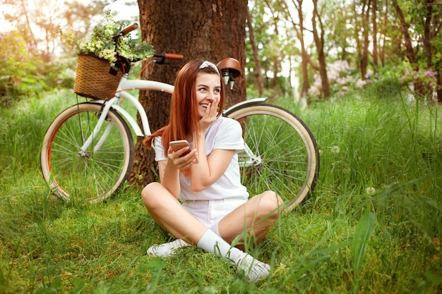 Vrouw rust in de natuur. Een meisje zit op het gras met haar telefoon. Fiets te huur en dagverhuur. Zomer lente. Technologie, internet om te communiceren met vrienden op vakantie, sociale netwerken. Glimlachend