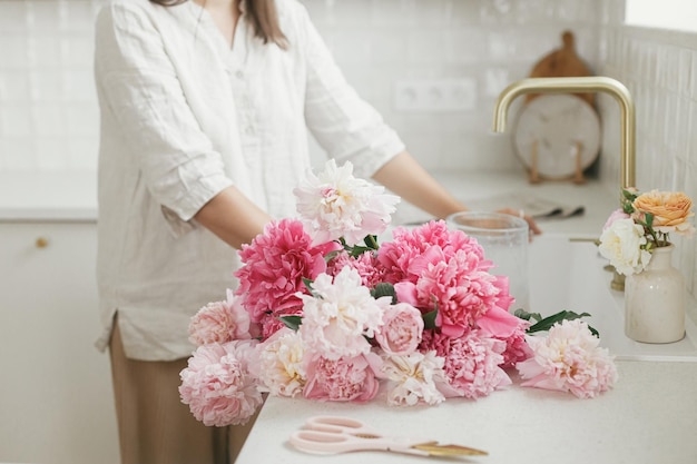 Vrouw schikken van mooie pioenrozen in vaas bij gootsteen met koperen kraan in nieuwe moderne keuken Vrouw versieren van bloemen op achtergrond van granieten aanrecht handen met bloemen close-up