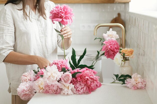 Vrouw schikken van mooie pioenrozen in vaas bij gootsteen met koperen kraan in nieuwe moderne keuken Vrouw versieren van bloemen op achtergrond van granieten aanrecht handen met bloemen close-up