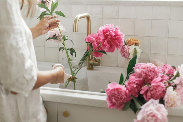 Vrouw schikken van mooie pioenrozen in vaas bij gootsteen met koperen kraan in nieuwe moderne keuken Vrouw versieren van bloemen op achtergrond van granieten aanrecht handen met bloemen close-up