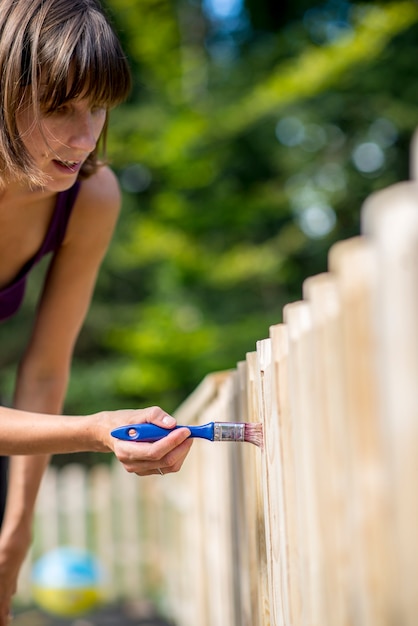 Vrouw schildert een houten houten tuinhek met een kleine kwast terwijl ze tuinonderhoud doet.