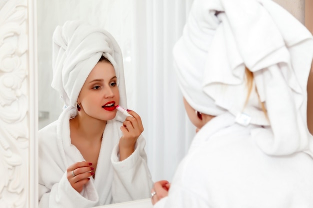 Vrouw schildert haar lippen in de badkamer van het hotel na het douchen. Mooie vrouw van Slavische verschijning in kamerjas over spiegel. Gelukkige dame. Gezonde gezichtshuid. Schattig meisje waste haar gezicht en toont emoties