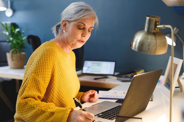 Vrouw schrijft in een notitieboek in een modern thuiskantoor met behulp van een laptop met een bureaublamp en een potplant