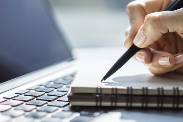 Foto vrouw schrijft met een pen in een notitieblok op het toetsenbord van de laptop in een zonnig kantoorconcept voor zaken en onderwijs close-up