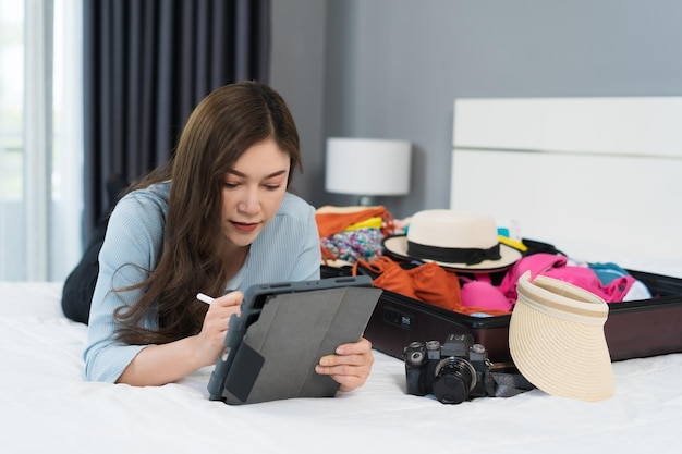 Vrouw schrijft tablet en verpakt kleding in koffer op een bed thuis reisvakantie plannen