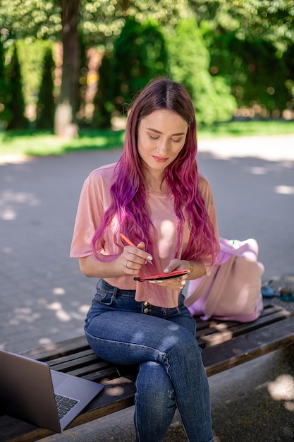 Vrouw schrijven in een notitieboekje zittend op een houten bankje in het park meisje buitenshuis werken op draagbare ...