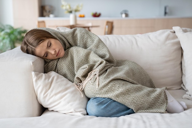 Vrouw slaapt thuis op de bank, rust in een plaid en voelt zich slecht. Symptomen griep covid seizoen.