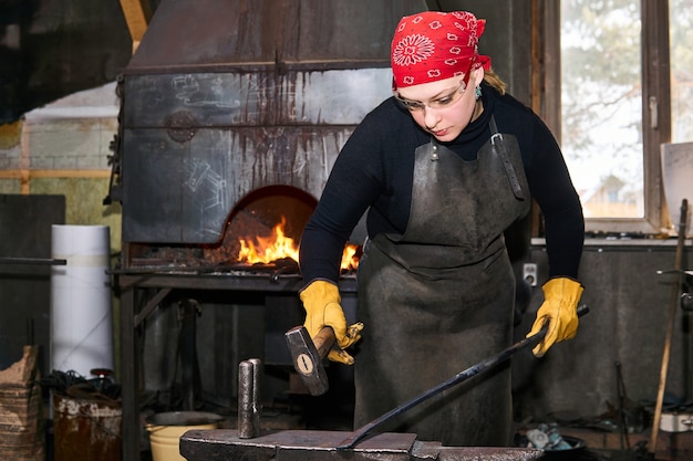 Vrouw smid metaalkunstenaar smeedt een heet metalen werkstuk met hamer op het aambeeld in een traditionele werkplaats