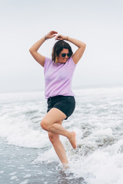 Vrouw speelt met zeewater heeft plezier, voelt zich vrij, draagt een zonnebril, een handgebreide blouse en een korte broek. Kopieer de ruimte Strand leuk concept