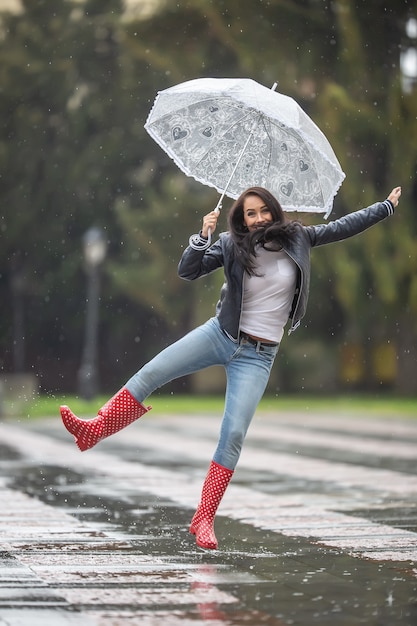 Vrouw springt van vreugde op een regenachtige dag met een paraplu en rode polka dot regenlaarzen.