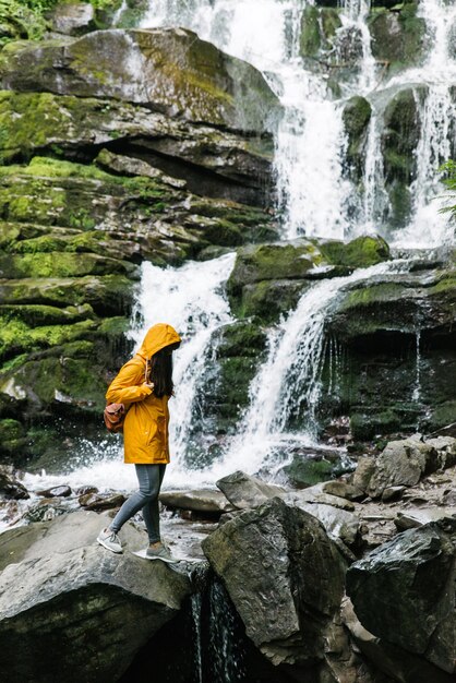 Vrouw staande op rots in gele regenjas met waterval op achtergrond