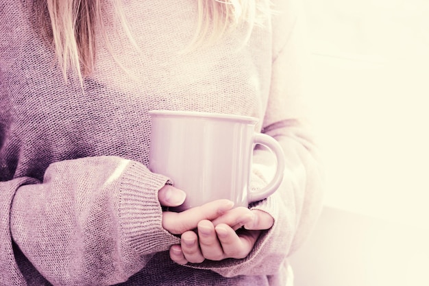 Vrouw staat bij het raam en houdt een kopje warme drank vast in de huiselijke sfeer Het lentezonlicht stroomt uit het raam