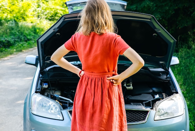 Vrouw staat in de buurt van de kapotte auto in de natuur. Ongevalsituatie tijdens het reizen. Meisje in jurk weet niet wat te doen. Open de motorkap van het voertuig.