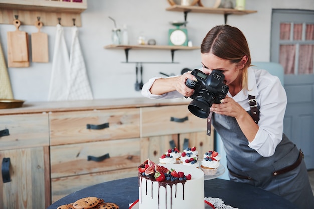 Vrouw staat in de keuken en neemt foto van haar zelfgemaakte koekjes en taart met behulp van camera.