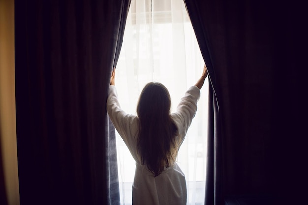 Vrouw staat in een badjas bij het raam en opent het gordijn in het hotel