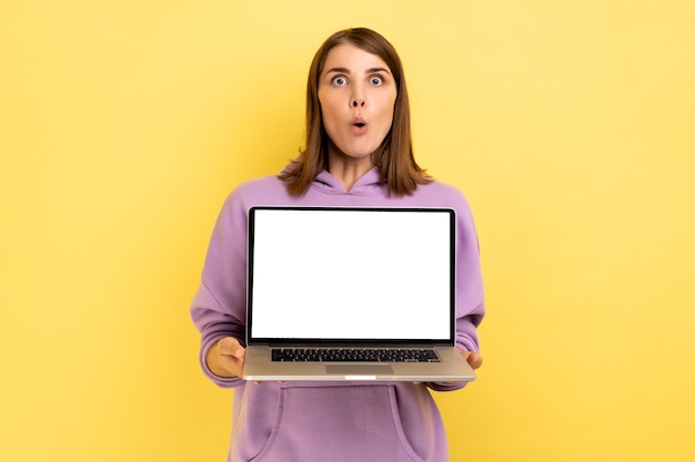 Vrouw staat met laptop met wit leeg scherm voor promotie en kijkt naar camera met open mond