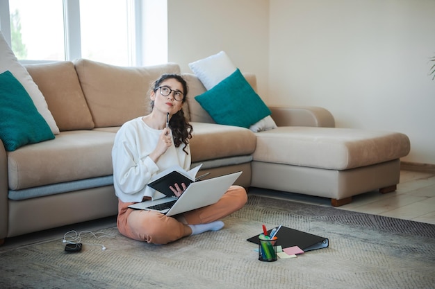 Vrouw studeert thuis met gekruiste benen op de vloer, leunend tegen een bank werkend met een laptop en...