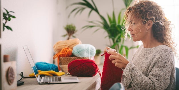 Vrouw thuis in vrijetijdsbesteding leren van online klas om werk te breien Vrouwelijke mensen die laptop gebruiken voor haar hobby of alternatief werk Dame van middelbare leeftijd breien met rode wol Concept van het gebruik van web