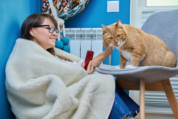 Vrouw thuis met behulp van smartphone opwarming met kat in de buurt van verwarmingsradiator