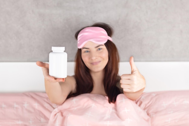 Foto vrouw toont een witte pot voor een goede slaap slapeloosheid pillen mock up een mooi meisje in bed in roze slaap masker ze sliep goed en was gelukkig toont duimen omhoog roze beddengoed