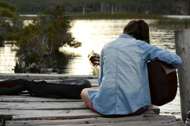 vrouw van achteren die gitaar speelt aan de oever van een meer op een pier