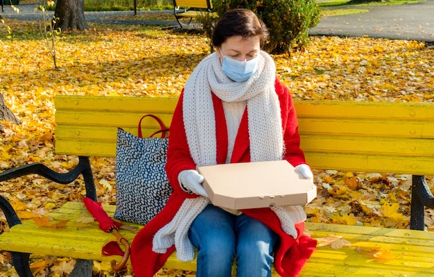 Vrouw van middelbare leeftijd 50 plus met beschermend medisch masker zittend op een gele bank in herfst park met doos voor pizza.