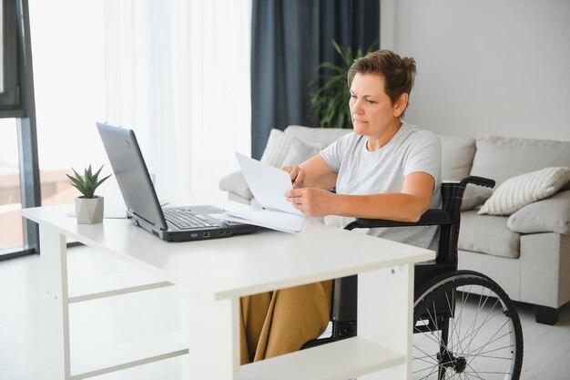 Vrouw van middelbare leeftijd die een laptop gebruikt die thuis op een rolstoel zit